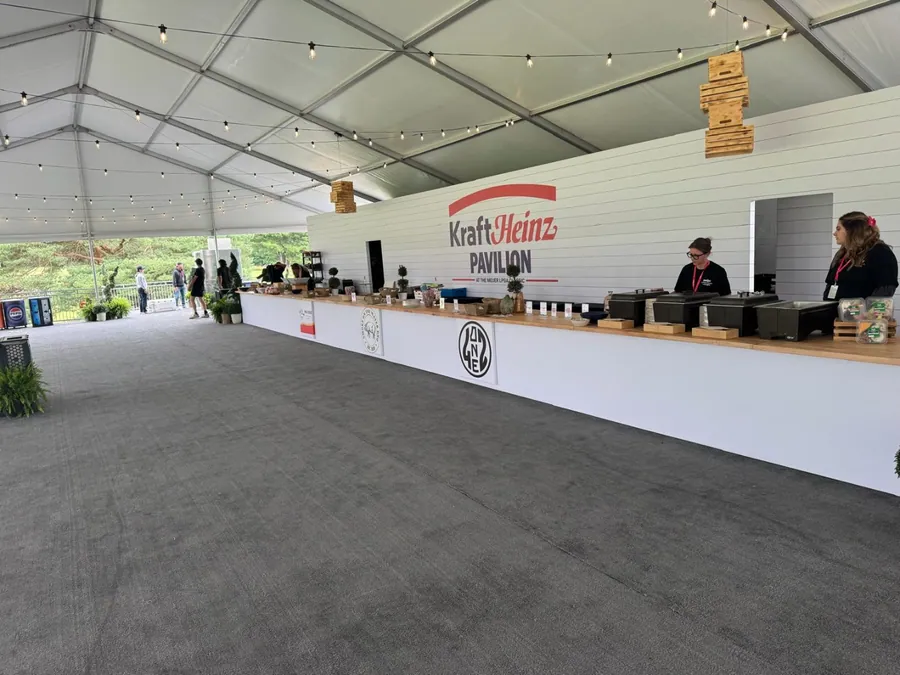 A long custom-built service counter with a branded back wall runs the length of a clear-span event pavilion, with staff working behind it and greenery at the entrance.
