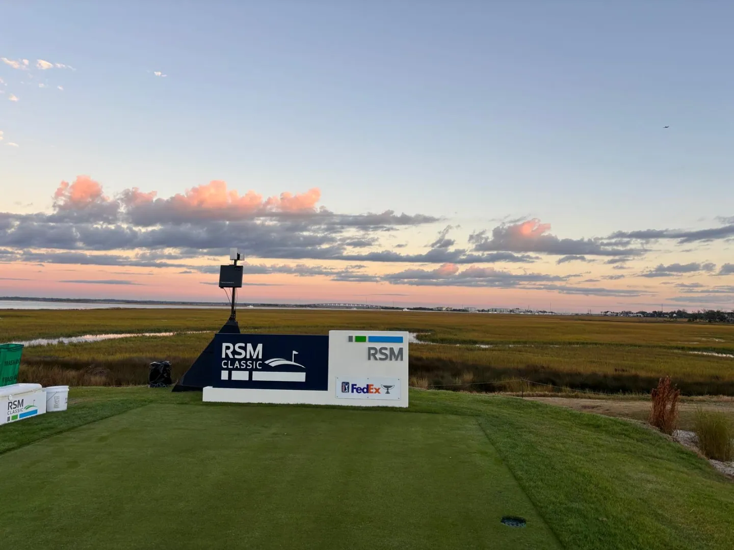 A tee box dressed with branded panels and a marker at dusk, looking out over a coastal course under a pink and blue sky.