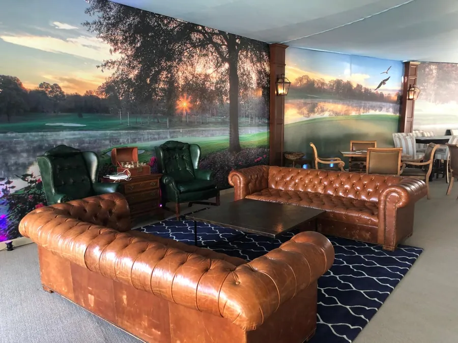 A lounge with tan leather chesterfield sofas and green wing chairs on a patterned rug, against a wall-length printed sunrise mural.