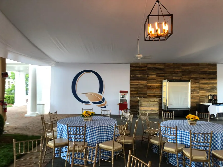Round tables in blue checked linen with sunflower centrepieces under a lantern chandelier, beside a timber-clad wall with an oversized graphic.