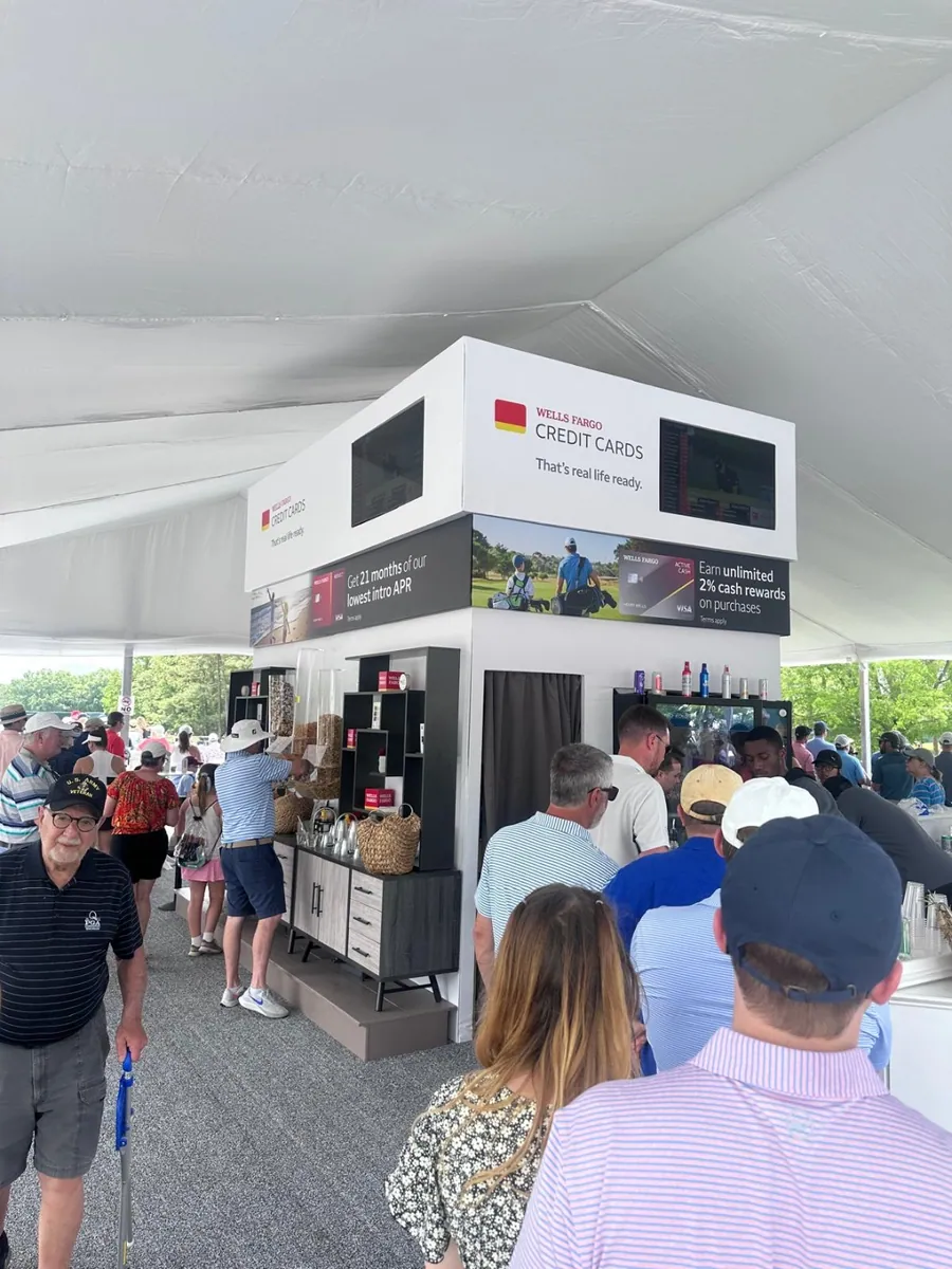 Spectators queueing at a white branded concession structure with a canopy roof on a course during an event.