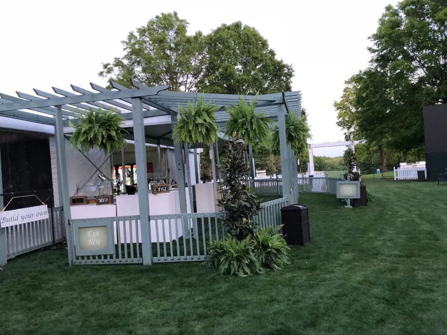 A white pergola structure planted with palms and hedging, enclosed by picket fencing on a lawn beside event tents.