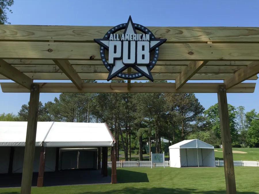 A timber pergola structure carrying a large circular branded pub sign, standing on a course in front of white event tents.